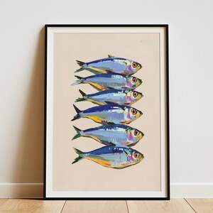 May include: A framed art print featuring a vertical stack of six stylised fish. Each fish is rendered in shades of blue, silver, and yellow, set against a solid light beige background. The frame is thin and black, enclosing the entire illustration.