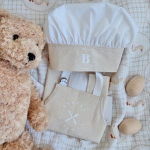 May include: A beige chef's hat and apron set with white details. The hat features a beige band with a white embroidered letter "B". The apron has the text "BROOKS KITCHEN" and a fork and knife graphic. Two wooden eggs and a teddy bear are also in the image.