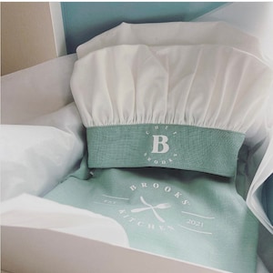 May include: A white chef hat with a green band that says "Chef Brooks" and a white letter "B" in the center. The hat is in a white box with a green linen fabric with the text "Brooks Kitchen Est. 2021" in white.