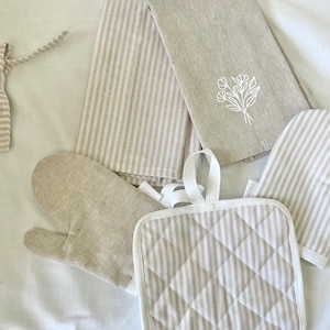 May include: A collection of kitchen linens in neutral tones. Includes striped oven mitts, a pot holder, and a tea towel with a floral embroidery. The set is designed with a farmhouse aesthetic.