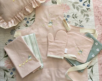 May include: A pink kitchen apron with a duck and flower embroidery design, a matching pink oven mitt, and two pink and green tea towels with duck embroidery.