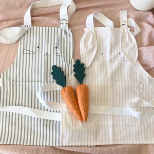 May include: Two striped bunny aprons with long ears, one in white and pink stripes and one in blue and white stripes. Two orange felt carrots with green felt leaves are in front of the aprons.