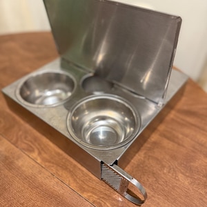 May include: A stainless steel food warmer with two round bowls and a hinged lid. The warmer has a rectangular base and a small grater with a handle. The unit is designed to keep food warm.