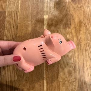 May include: A pink plastic piggy bank with a small black eye and a slot on top. The toy pig has a small opening on the side and a pink snout. The piggy bank is on a wooden surface.
