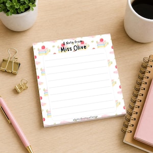 以下が含まれることがあります： 「A Note from Miss Olive」のテキストと本とリンゴの装飾パターンが施された、罫線入りのメモ帳。 メモ帳は木製の表面に置かれ、ピンクのペン、金色のペーパークリップ、白い鉢植えの小さな植物があります。
