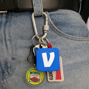 May include: A collection of keys and keyrings attached to a silver carabiner clipped to a belt loop. The keyrings include a blue square with a white "V" logo, a round keyring with text and a strawberry graphic, and a rectangular tag.
