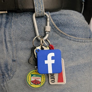 May include: A set of keyrings suspended from a silver carabiner attached to a belt loop. The keyrings feature a blue square with a white "f" logo, a round yellow tag with a strawberry image, and a rectangular tag with red and white stripes.