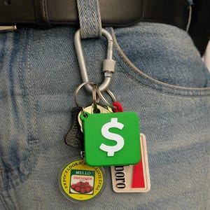 May include: A set of keys hanging from a silver carabiner attached to a black belt. The keys include a green square keyring with a white dollar symbol, a round yellow keyring with text, and a rectangular keyring with red details.