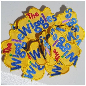 May include: A pile of yellow felt patches with the words "The Wiggles" embroidered in blue, purple, and red. The patches are in various shapes and sizes.