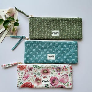 Puede incluir: Tres estuches rectangulares con cremallera en tonos verde, turquesa y estampado floral. El estuche verde tiene un pequeño diseño de hojas, el turquesa tiene un diseño de lazo y el floral tiene flores rosas y rojas. Cada estuche tiene una correa de muñeca y una etiqueta blanca que dice "l'étoffe".