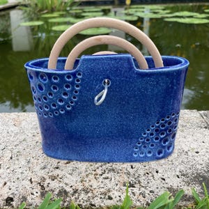 Puede incluir: Una cesta de cerámica azul cobalto con dos asas de madera. La cesta tiene una superficie texturizada y recortes circulares decorativos. Un gancho de metal plateado está unido a la parte delantera. La cesta está sobre una superficie de piedra.