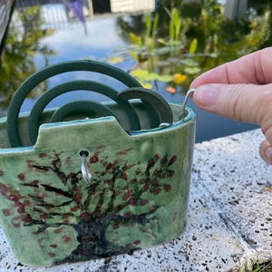 Puede incluir: Un soporte de cerámica verde con un diseño de árbol, que contiene varias espirales repelentes de mosquitos de color verde oscuro. El soporte se coloca cerca de un estanque con reflejos de plantas y cielo.