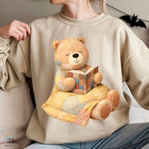 Peut inclure: Sweat-shirt beige avec une illustration à l'aquarelle d'un ours en peluche patchwork lisant un livre. L'ours est assis sur une couverture patchwork jaune. Le sweat-shirt a un col rond et des manches longues. Le texte "Chill Chill Digital" est en bas à gauche.