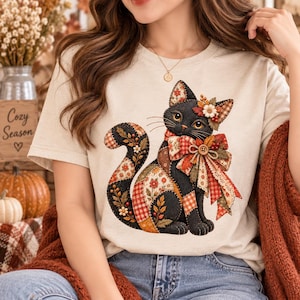 May include: A beige short-sleeve t-shirt featuring a graphic of a black cat with a patchwork-style body. The cat is decorated with floral and plaid patterns, a large plaid bow around its neck with a button center, and autumn-themed floral accents.