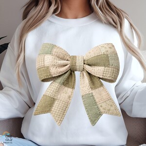 May include: White sweatshirt featuring a large, patchwork bow design. The bow is composed of various fabric squares in shades of green, beige, and cream, with a quilted texture. The bow is the focal point of the shirt.