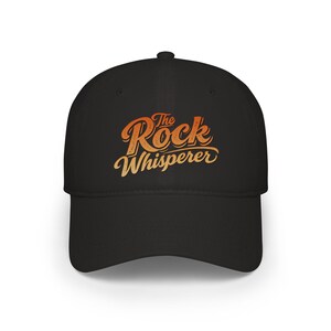 May include: Black baseball cap with the text "The Rock Whisperer" in orange and yellow script. The cap has a curved brim and a structured crown. A stylish accessory for outdoor activities.