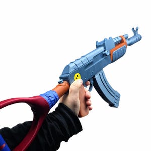 May include: A blue and brown toy rifle featuring a yellow smiley face sticker on the side. The stock is connected to a red curved handle with blue tape wrapped around the joint. The toy has a detachable magazine and a front sight post.