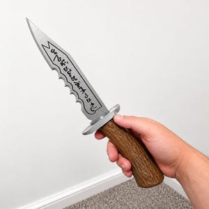 May include: A silver-colored knife with a serrated edge and a brown, textured handle that resembles wood grain. The blade features a decorative banner design containing black, stylized, non-Latin script characters running along its length.
