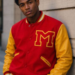 May include: A red varsity jacket featuring yellow sleeves, a large yellow letter M on the chest, and a snap-button front closure. The collar, cuffs, and hem are ribbed with red, white, and yellow stripes. The jacket includes two side pockets.