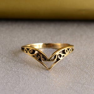 Könnte beinhalten: Ein goldfarbener Ring mit V-förmigem Design. Der Ring hat ein durchbrochenes Muster mit wirbelnden Details am oberen Rand. Der Ring ist vor einem neutralen Hintergrund platziert.