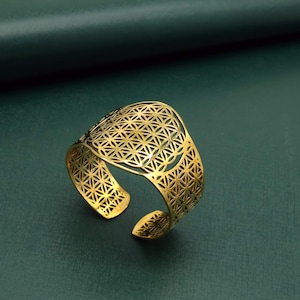 May include: A gold-colored metal cuff bracelet featuring an intricate, repeating geometric pattern of overlapping circles known as the flower of life. The bracelet has an open-ended design, allowing for adjustment, and displays a polished finish.