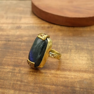 Könnte beinhalten: Goldfarbener Ring mit einem großen, rechteckigen, dunkelblauen Edelstein. Der Ring hat dekorative goldfarbene Akzente an der Lünette und am Band. Der Edelstein hat eine schillernde Qualität, auf einer Holzoberfläche.