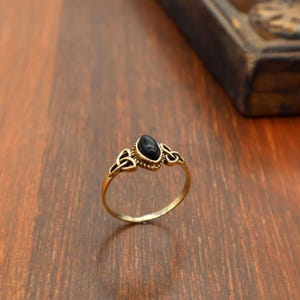 Può includere: Un anello dorato con una pietra nera di forma ovale. L'anello presenta un design decorativo sulla fascia con dettagli a nodo celtico. Appoggiato su una superficie di legno.