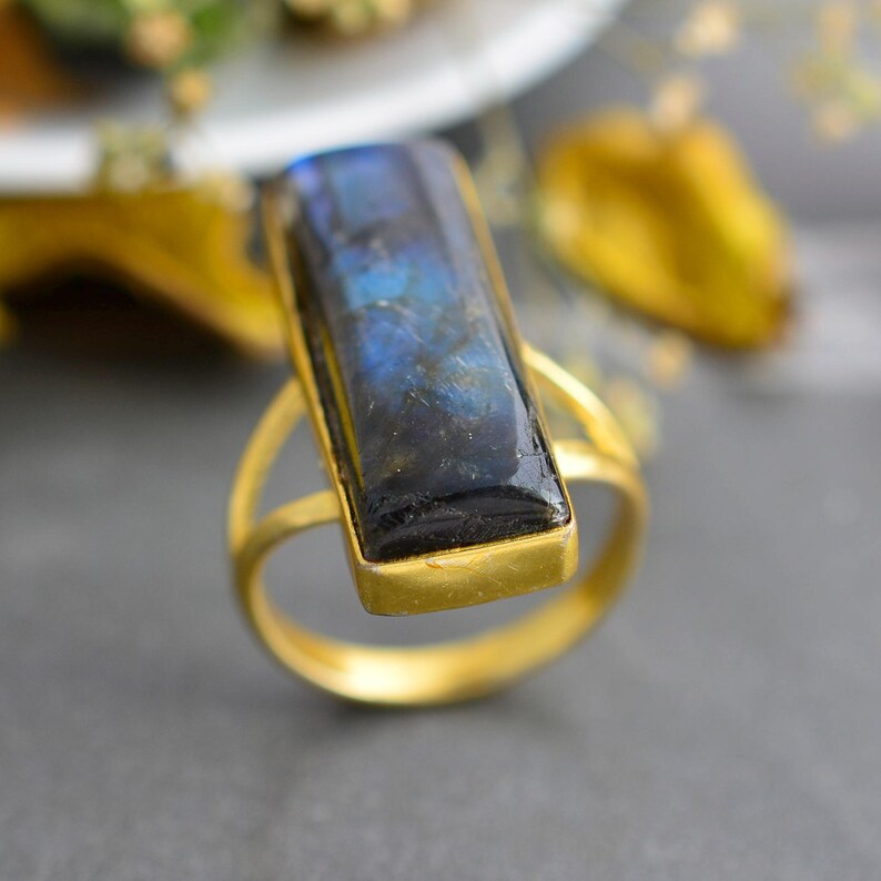 May include: A gold-toned ring featuring a rectangular, elongated labradorite gemstone set in a smooth metal bezel. The stone displays deep blue and dark grey iridescent flashes across its surface, mounted on a simple, thin, circular gold-coloured band.