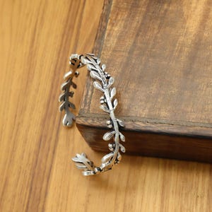 Könnte beinhalten: Ein silberfarbenes Armreif-Armband mit Blattdesign. Das offene Armband weist detaillierte Blätter und kleine runde Akzente auf. Das Armband wird auf einer Holzoberfläche präsentiert, die sein kompliziertes Design und seine metallische Oberfläche zeigt.