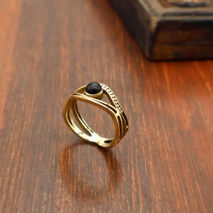 May include: A gold-coloured ring featuring a black stone at its centre. The ring has a layered design with a dotted pattern along the top band. The ring is displayed on a wooden surface.