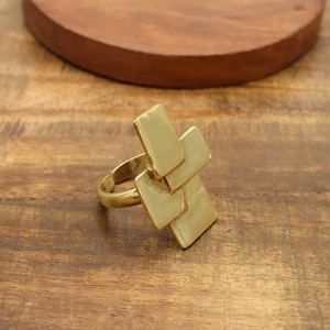 May include: A gold-colored ring with a geometric design. The ring features a series of overlapping rectangular shapes, creating a modern and artistic look. The ring is set against a wooden background.