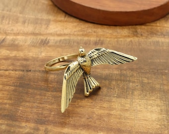 May include: A gold-coloured metal napkin ring shaped like a bird with spread wings. The bird has feather detailing and is attached to a circular ring. The napkin ring sits on a wooden surface.
