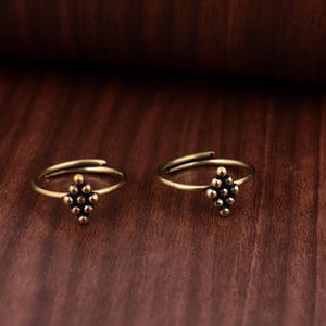 May include: Two gold-colored toe rings with a cluster of small, dark-colored beads at the center. The rings are set against a dark brown wooden surface, creating a contrast that highlights the jewelry's design. The rings are adjustable.