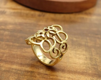 May include: A gold-colored ring with an openwork floral design. The ring features a series of interconnected loops and floral shapes, creating a decorative and ornate appearance. The ring is set against a wooden surface.