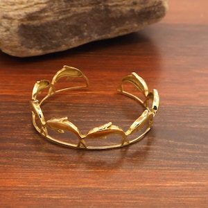 May include: A gold-colored cuff bracelet featuring a repeating dolphin design. The open-ended bracelet is made of a shiny, metallic material. The dolphins are arranged in a circular pattern around the wrist. The bracelet is displayed on a wooden surface.