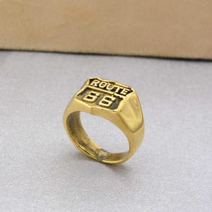 May include: A gold-colored ring with a rectangular top featuring the words "ROUTE 66" above the number "66". The ring has a classic signet style with a polished finish. The ring is a vintage-inspired accessory.