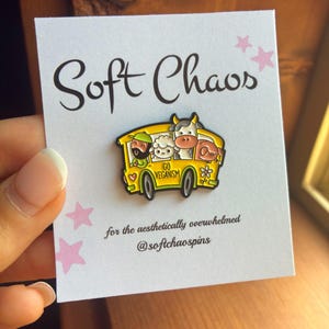 May include: A yellow enamel pin shaped like a school bus featuring a person, a sheep, a cow, and a pig. The bus has the text GO VEGANISM printed on the side. The pin is attached to a white display card with the text Soft Chaos and @softchaospins.