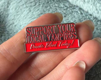 May include: A rectangular metal enamel pin with a silver border. The top section features the red text SUPPORT YOUR LOCAL VAMPIRES on a silver background. The bottom section is a solid red bar with the white text Donate Blood Today!