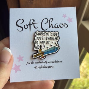 May include: A white enamel pin featuring a gold and blue sword with the black text 'CURRENT SIDE QUEST BROUGHT TO YOU BY ADHD' and small black hearts. The pin is attached to a white display card with the text 'Soft Chaos' and pink stars.