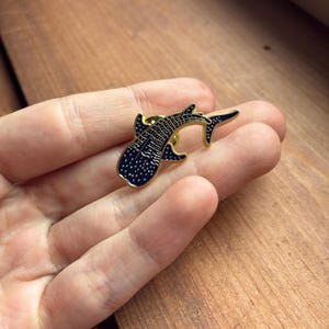 Pu&ograve; includere: Spilla in metallo color oro a forma di squalo balena. La spilla presenta un corpo blu scuro con pois bianchi e dettagli neri. Un piccolo accessorio, ideale per giacche, zaini o cappelli.