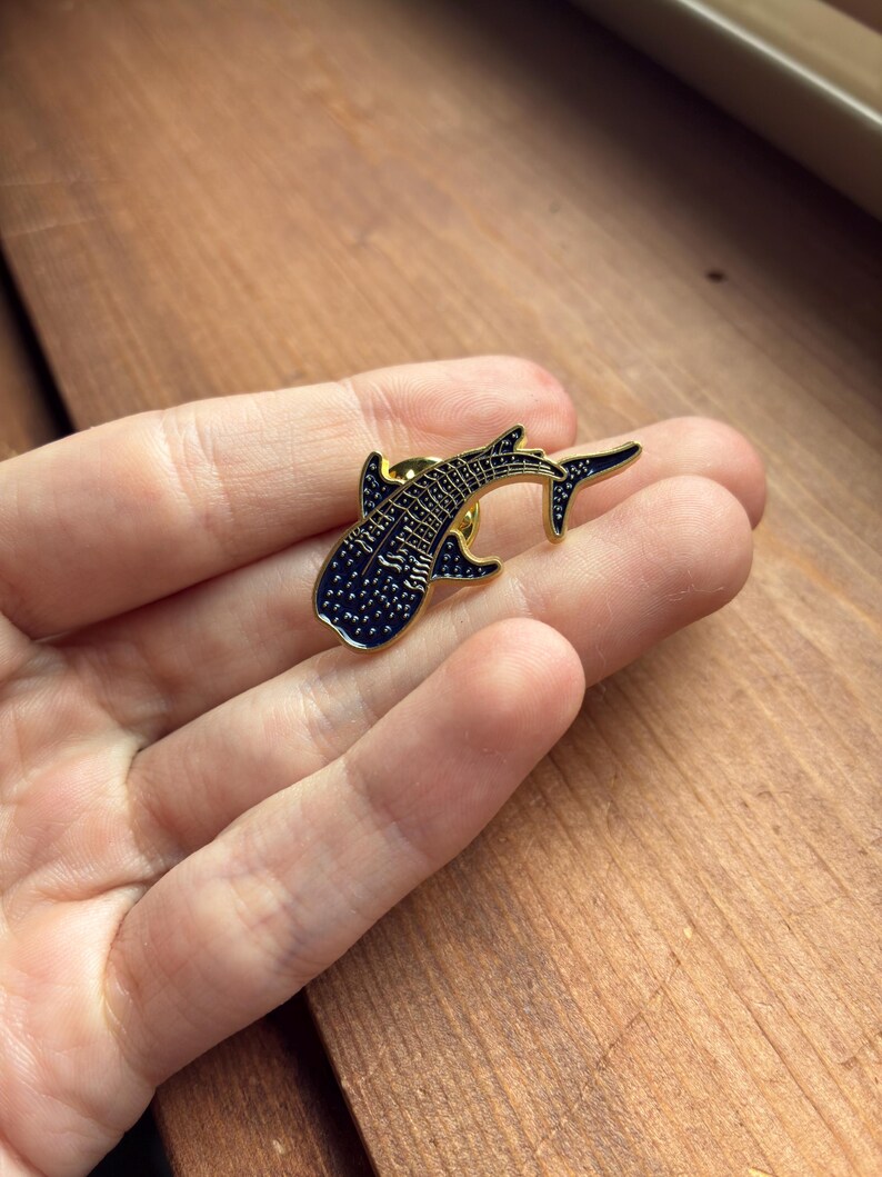 Pu&ograve; includere: Spilla in metallo color oro a forma di squalo balena. La spilla presenta un corpo blu scuro con pois bianchi e dettagli neri. Un piccolo accessorio, ideale per giacche, zaini o cappelli.
