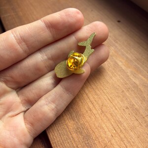 Pu&ograve; includere: Una spilla in smalto color oro a forma di balena. La spilla ha una chiusura color oro sul retro. La spilla &egrave; tenuta in mano su uno sfondo di legno. La spilla a forma di balena &egrave; un accessorio decorativo.