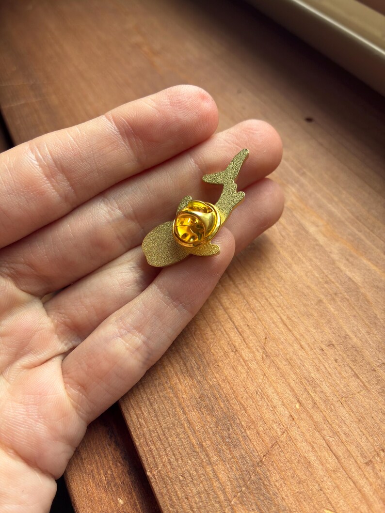 Pu&ograve; includere: Una spilla in smalto color oro a forma di balena. La spilla ha una chiusura color oro sul retro. La spilla &egrave; tenuta in mano su uno sfondo di legno. La spilla a forma di balena &egrave; un accessorio decorativo.