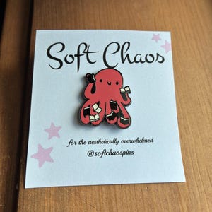 May include: A red octopus enamel pin featuring a smiling face and multiple arms holding small black and white books. The pin is attached to a white display card with the text Soft Chaos for the aesthetically overwhelmed @softchaospins.