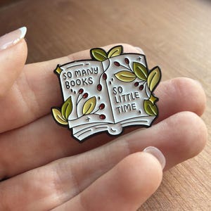May include: An enamel pin shaped like an open book with white pages and black outlines. The text reads SO MANY BOOKS SO LITTLE TIME. The design features green leaves and small red berries decorating the pages, accented with tiny gold star details.