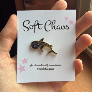 Pu&ograve; includere: Spilla in smalto a forma di squalo balena con contorno dorato e corpo blu navy, esposta su un cartoncino bianco. Il cartoncino presenta il testo "Soft Chaos" e il claim "for the aesthetically overwhelmed @softchaospins", insieme a illustrazioni di stelle rosa.