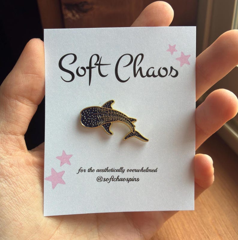Pu&ograve; includere: Spilla in smalto a forma di squalo balena con contorno dorato e corpo blu navy, esposta su un cartoncino bianco. Il cartoncino presenta il testo "Soft Chaos" e il claim "for the aesthetically overwhelmed @softchaospins", insieme a illustrazioni di stelle rosa.