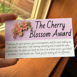 Pode incluir: Um cartão retangular com uma secção superior roxo-claro com um alfinete de esmalte dourado em forma de flor de cerejeira e o texto The Cherry Blossom Award. A secção inferior branca contém uma mensagem impressa de agradecimento.