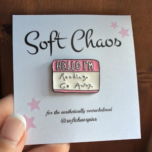 May include: A rectangular enamel pin featuring a pink and white name tag design with the black text 'HELLO I'M Reading. Go Away.' The pin is attached to a white display card printed with 'Soft Chaos', pink stars, and social media handle.