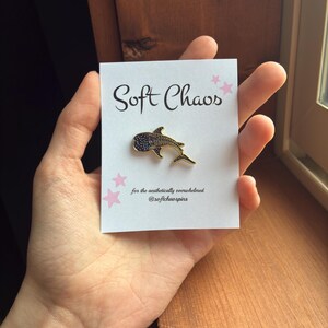 Pu&ograve; includere: Una spilla in smalto a forma di squalo balena dorato con dettagli neri &egrave; esposta su una carta bianca. La carta recita "Soft Chaos" in corsivo nero, con accenti di stelle rosa. &Egrave; presente anche il testo "for the aesthetically overwhelmed @softchaospins".
